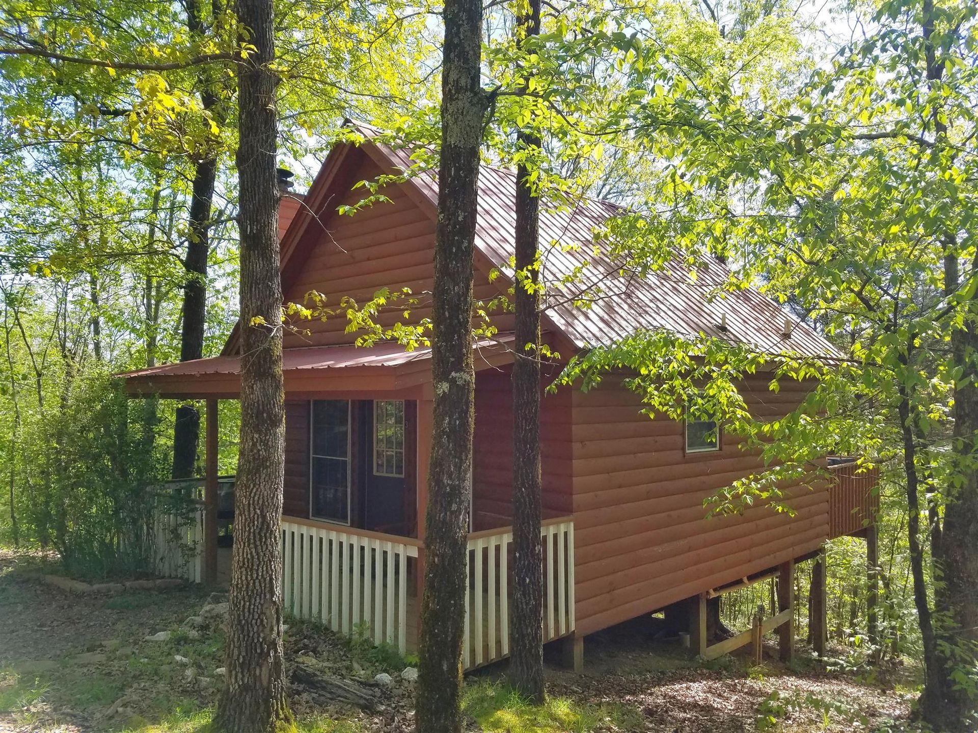 Small red cabin in a wooded area, with a porch and trees surrounding it.