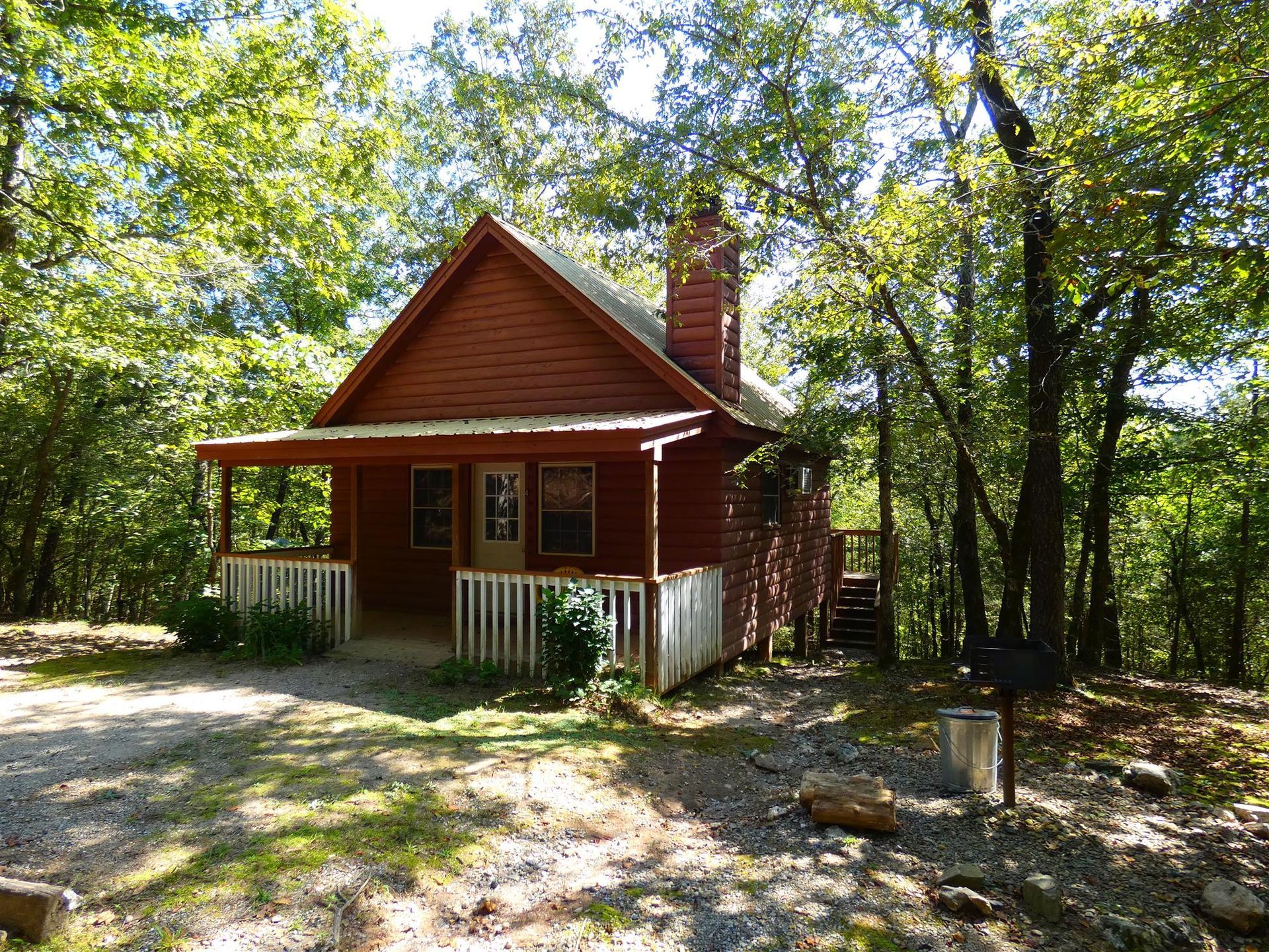 Small red cabin with a porch in a wooded area, surrounded by trees and sunlight.