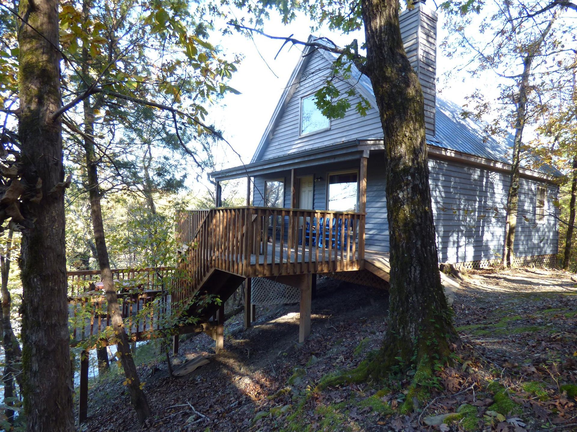 Wooded cabin with a deck and stairs, surrounded by trees on a sloped hillside.