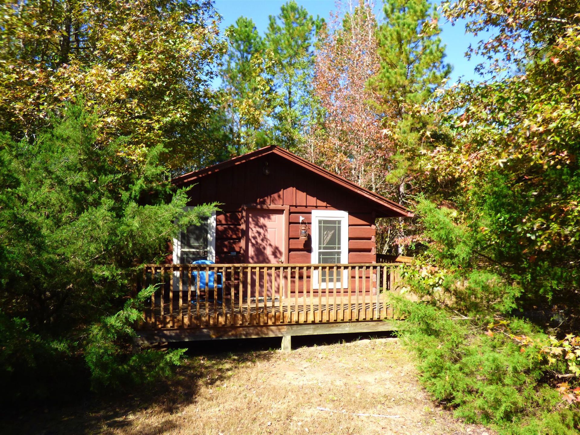 Red cabin with a wooden porch surrounded by dense green trees in a forest clearing.