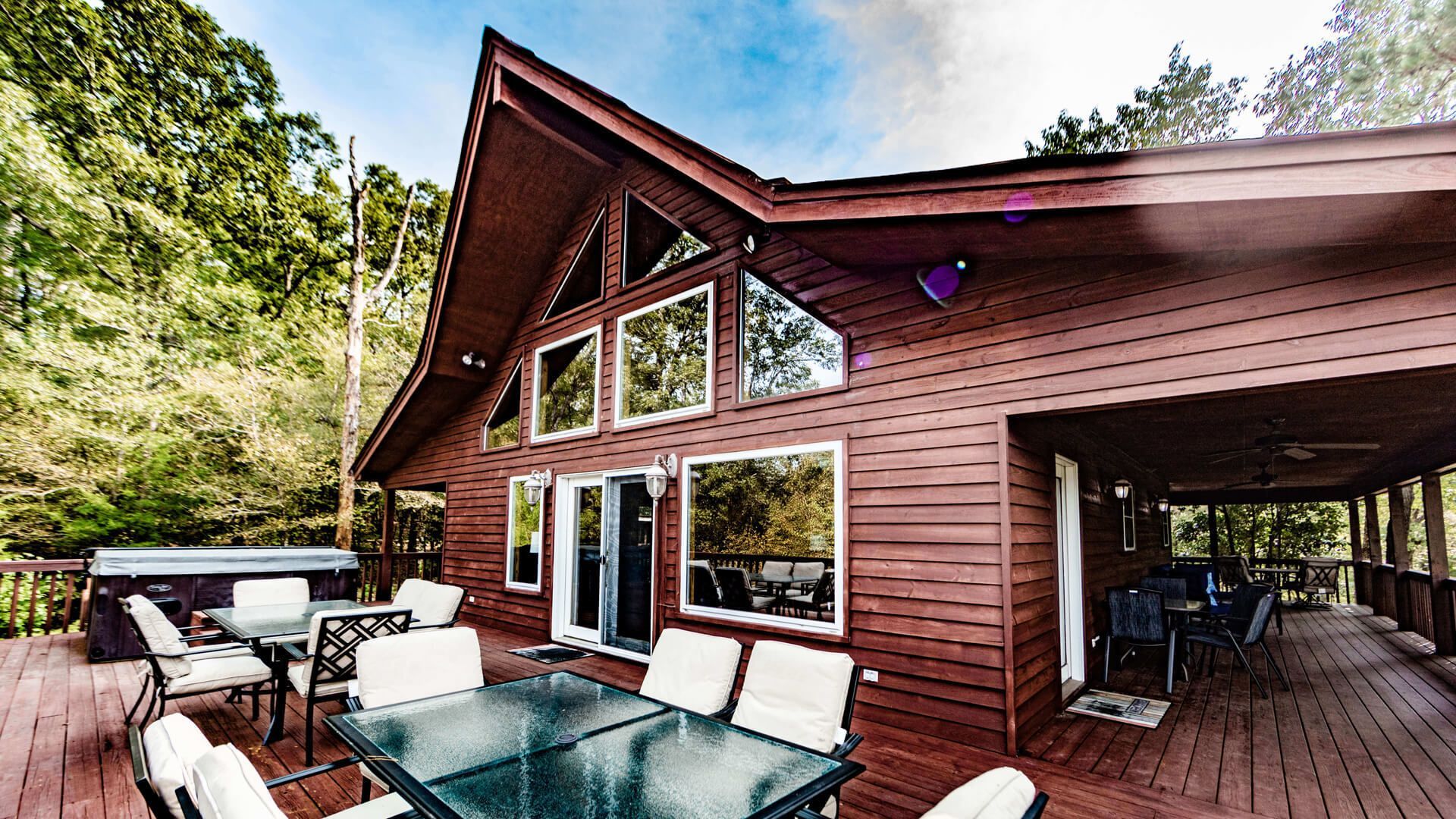 Wooden cabin deck with glass table and white patio chairs beneath a triangular roofline, surrounded by trees