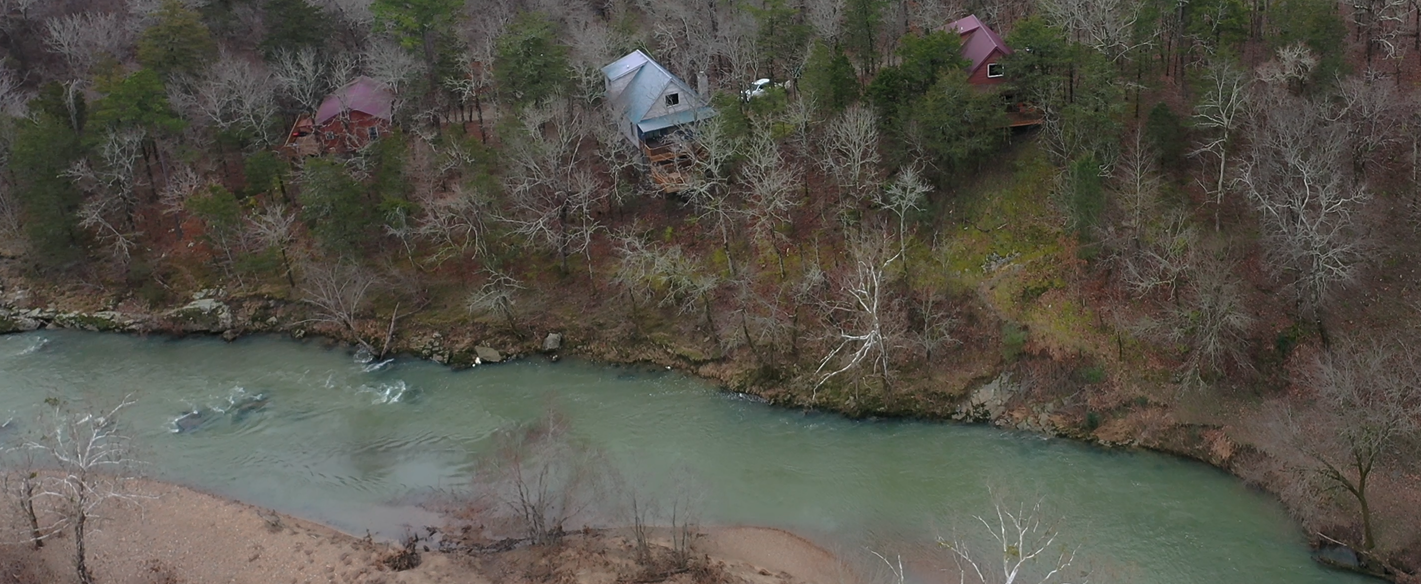 Aerial view of a narrow green river beside leafless trees and a small white tent on the bank