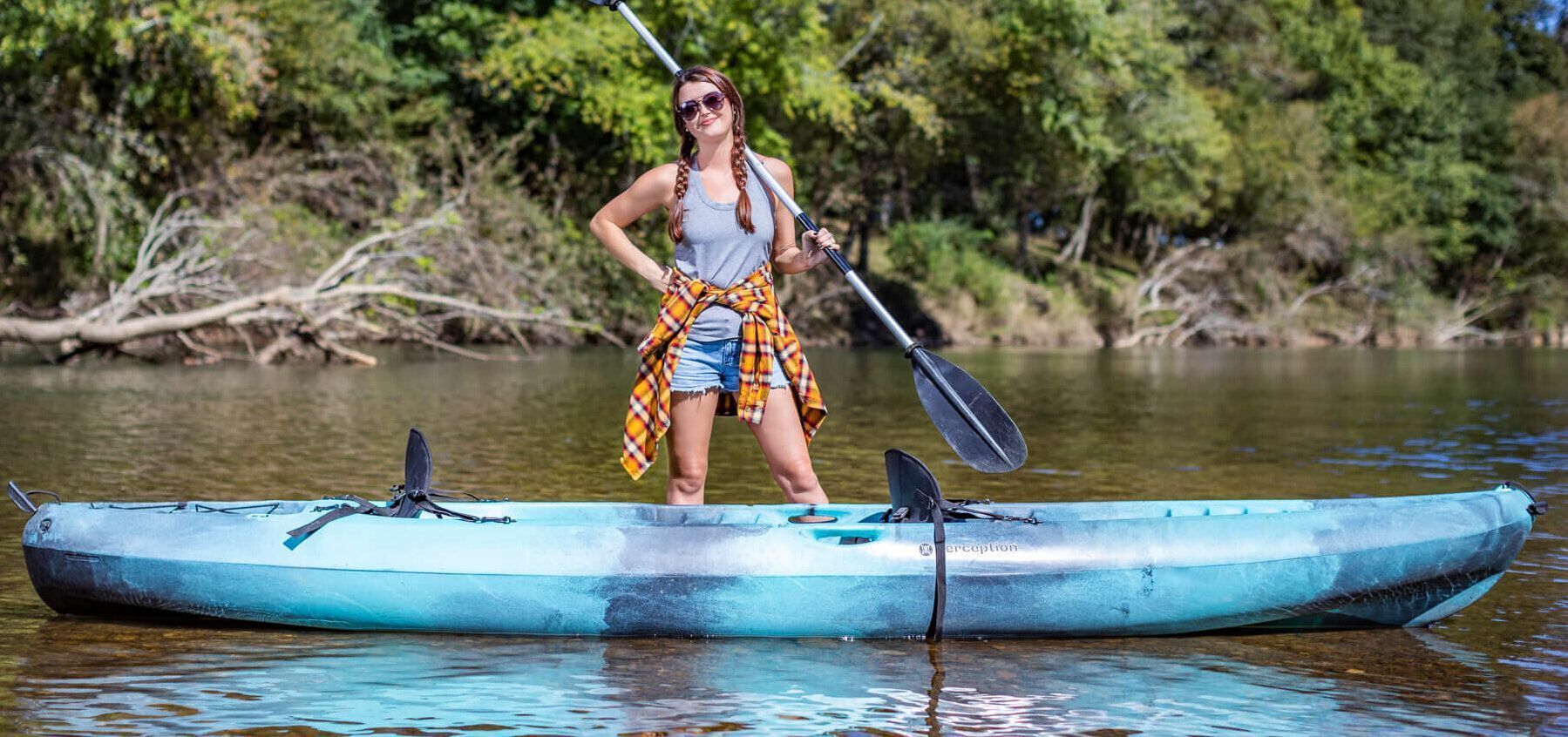 Person standing on a blue kayak with a paddle in calm water near a tree-lined shore