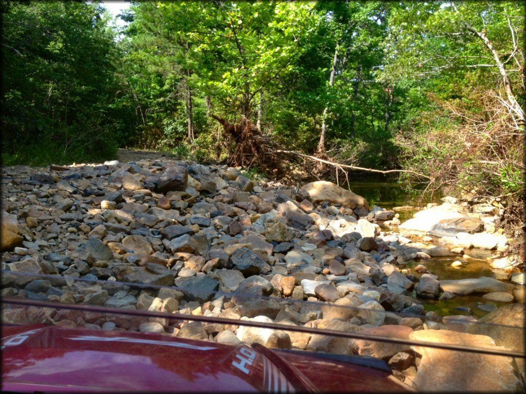 Rocky creek crossing in a wooded area, viewed over the hood of a red vehicle.