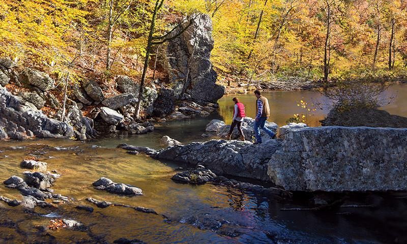 Two hikers on rocks beside a shallow river in a wooded autumn canyon