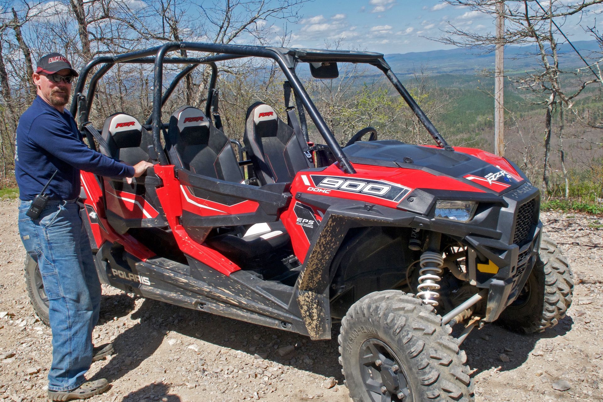 Man standing beside a red and black off-road side-by-side vehicle on a rocky mountain overlook
