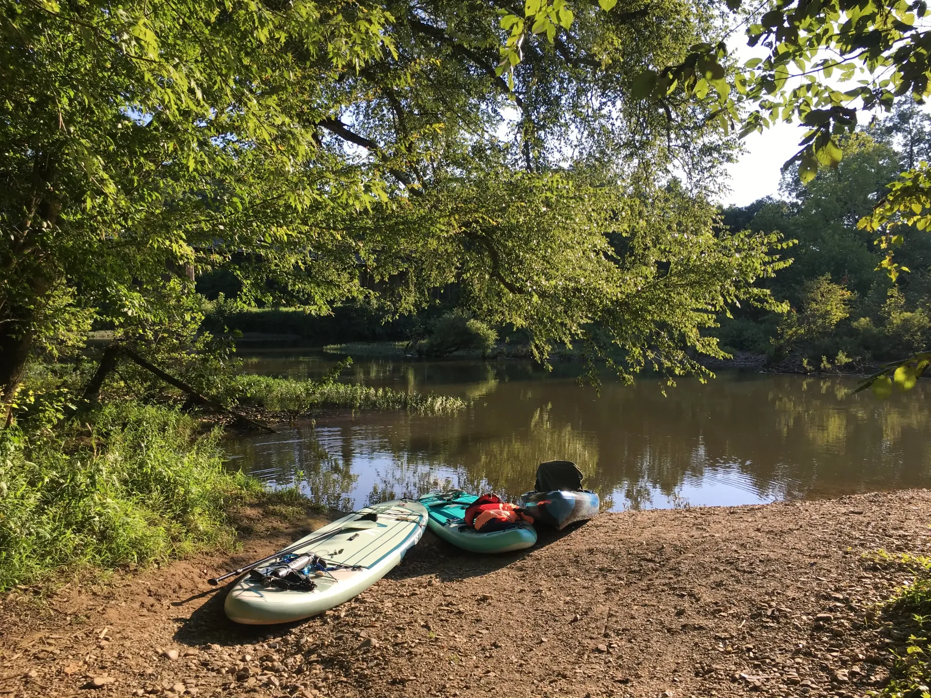 Two kayaks on a pebbled riverbank by calm water under leafy trees
