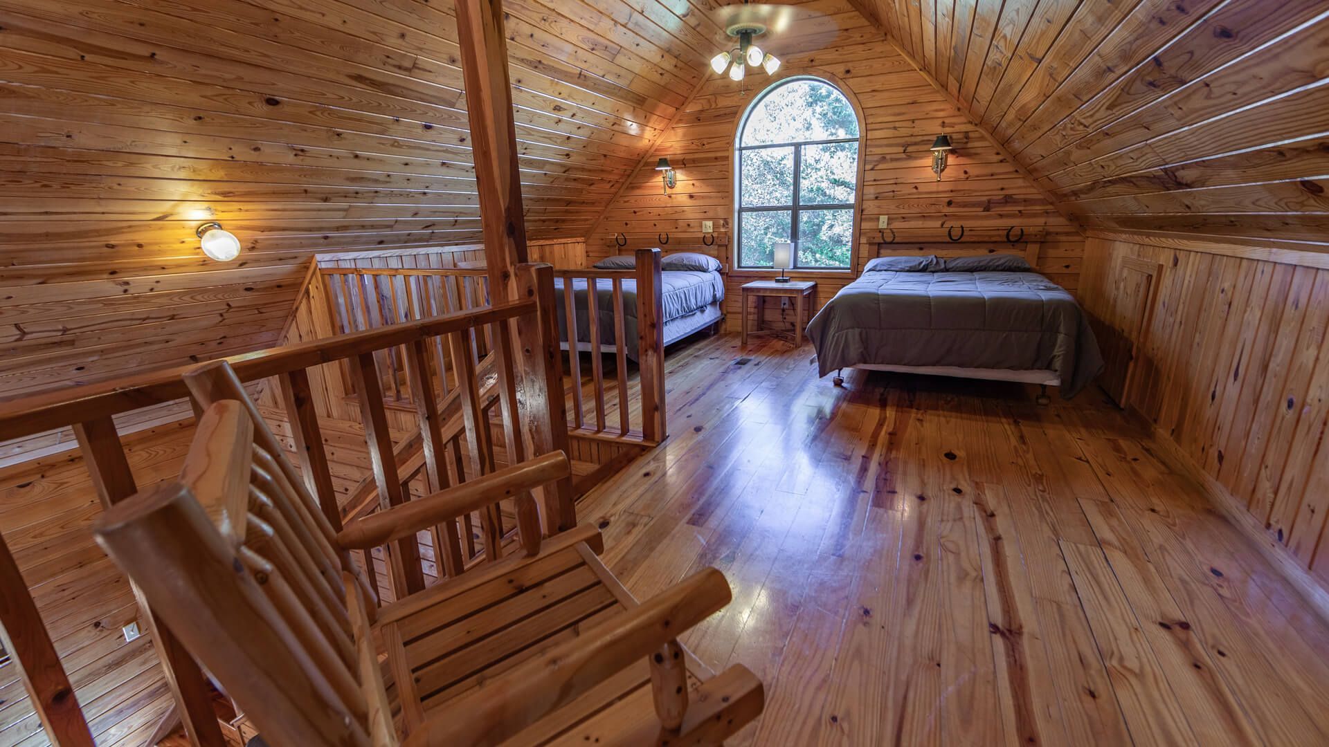 Cozy wood-paneled attic bedroom with a bed, rocking chair, and arched window overlooking trees