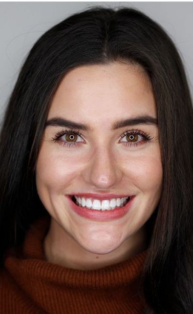 Smiling person with dark hair wearing a brown turtleneck against a plain gray background