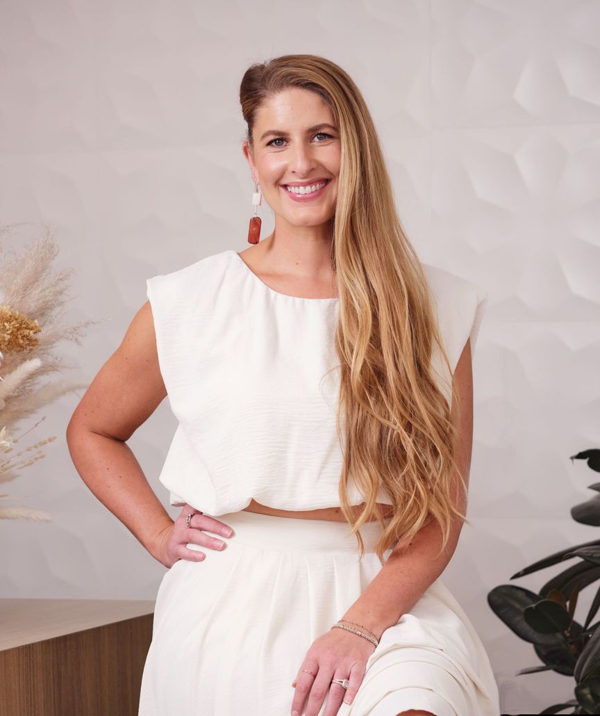 Smiling person in a white dress standing indoors against a light textured wall