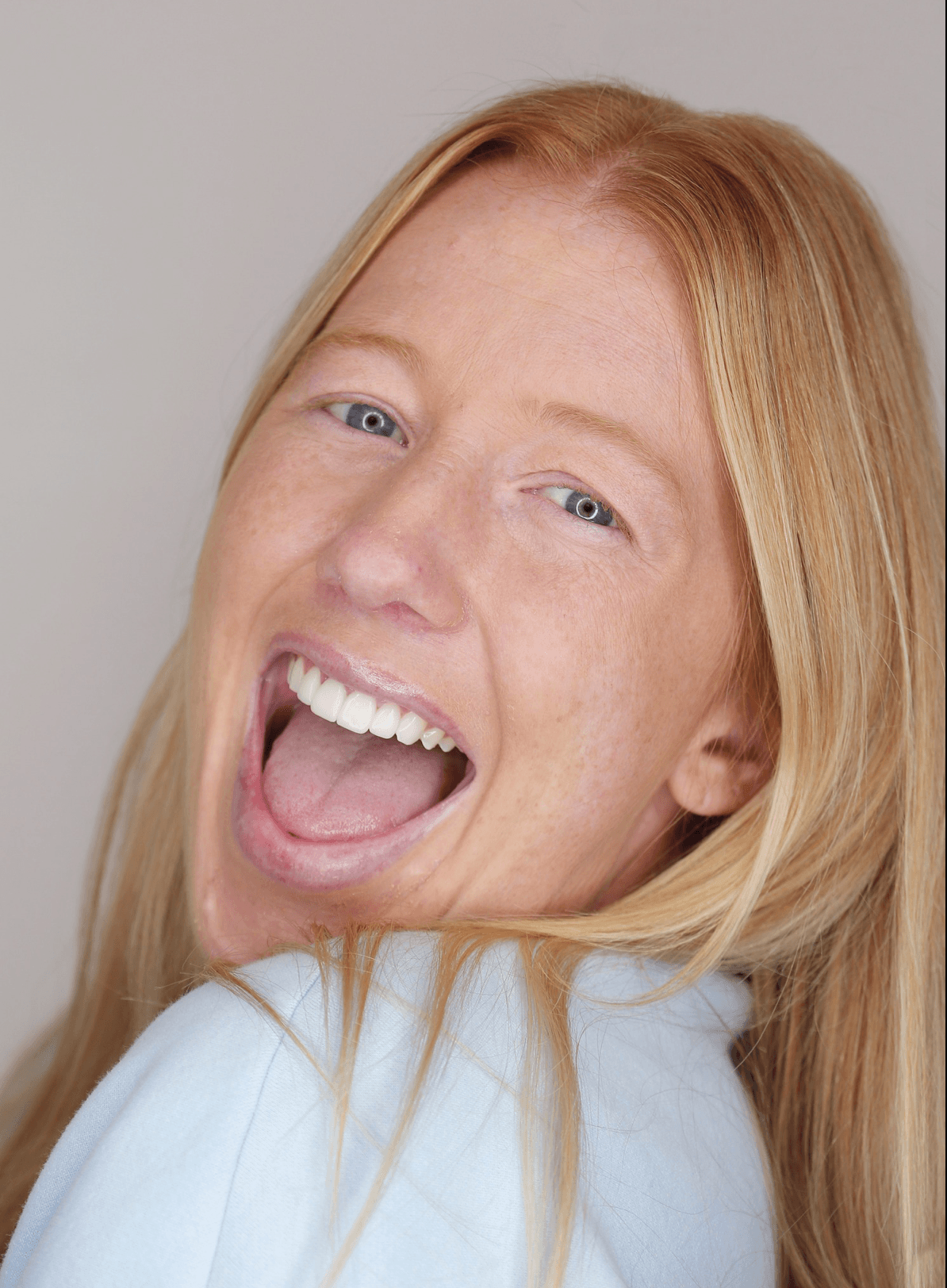 Smiling blonde woman with mouth open, wearing a light blue top against a gray background