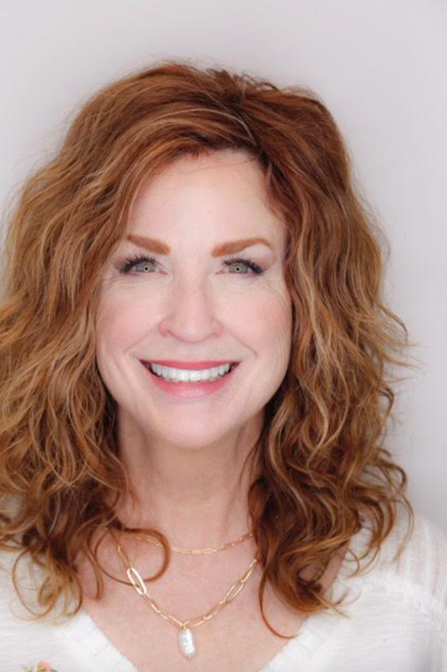 Smiling woman with curly red hair wearing a white top and gold necklace against a light background
