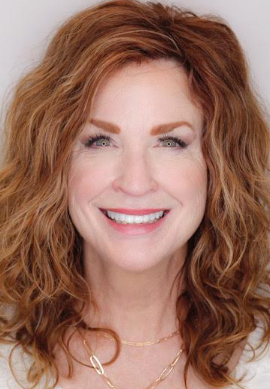 Smiling woman with curly red hair and a gold necklace, facing the camera on a light background