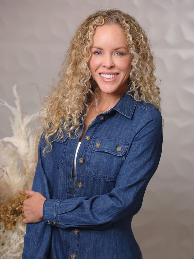 Smiling woman with long curly blonde hair wearing a brown off-shoulder top against a light background