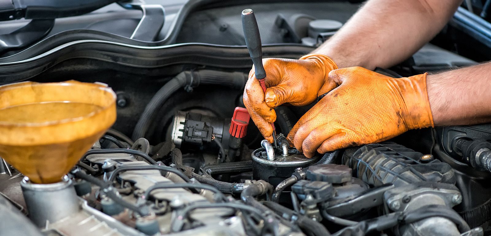 A mechanic wearing orange protective gloves uses a screwdriver while working on an open car engine with a yellow funnel.