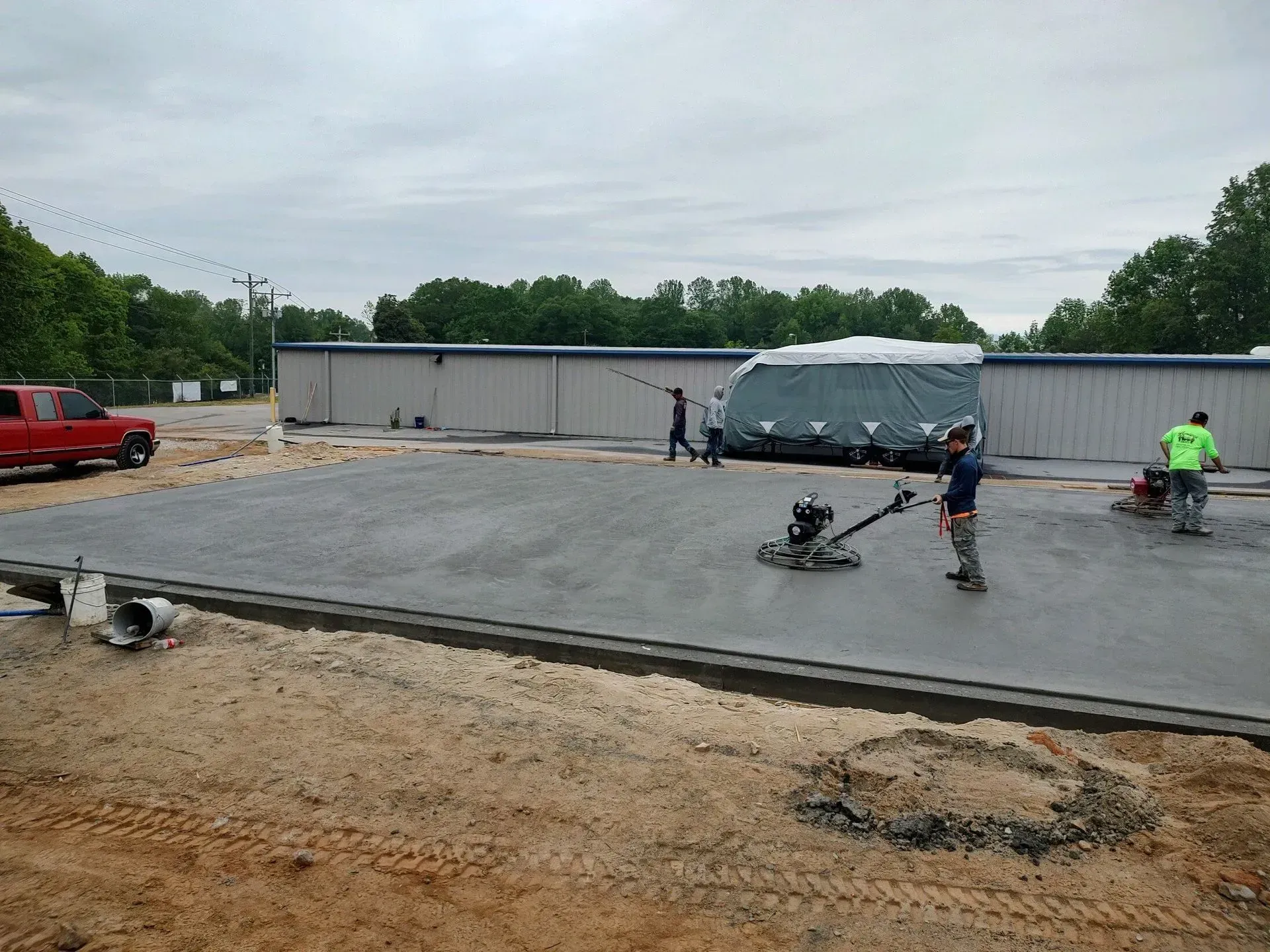 Workers finishing a concrete slab at a construction site with a white building and parked vehicles nearby