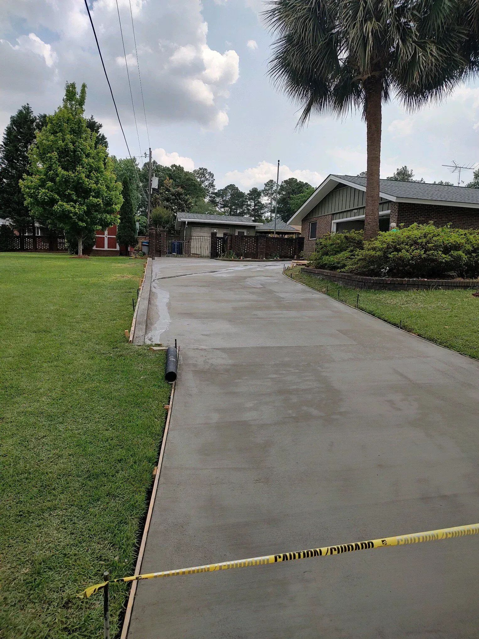 Concrete driveway leading to a house with a grassy lawn, palm tree, and yellow caution tape across the foreground.