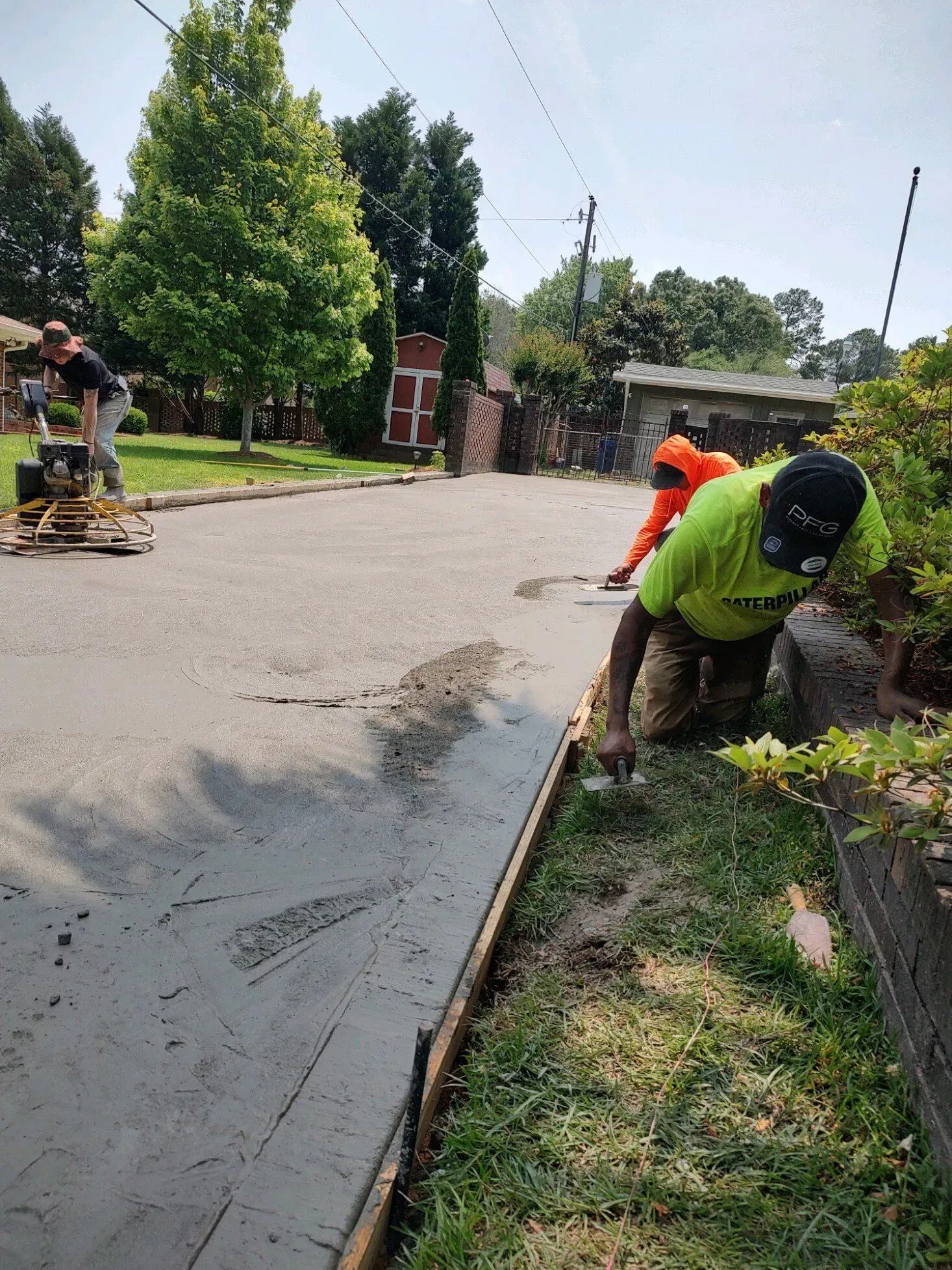 Worker in orange vest smoothing wet asphalt on a roadside, with trees and houses in the background.