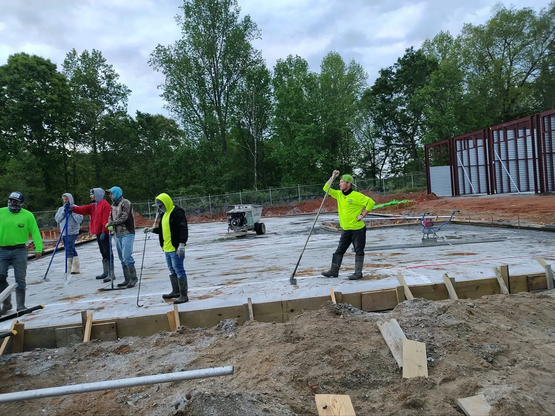 Construction workers in bright safety gear pour and smooth a concrete foundation outdoors.