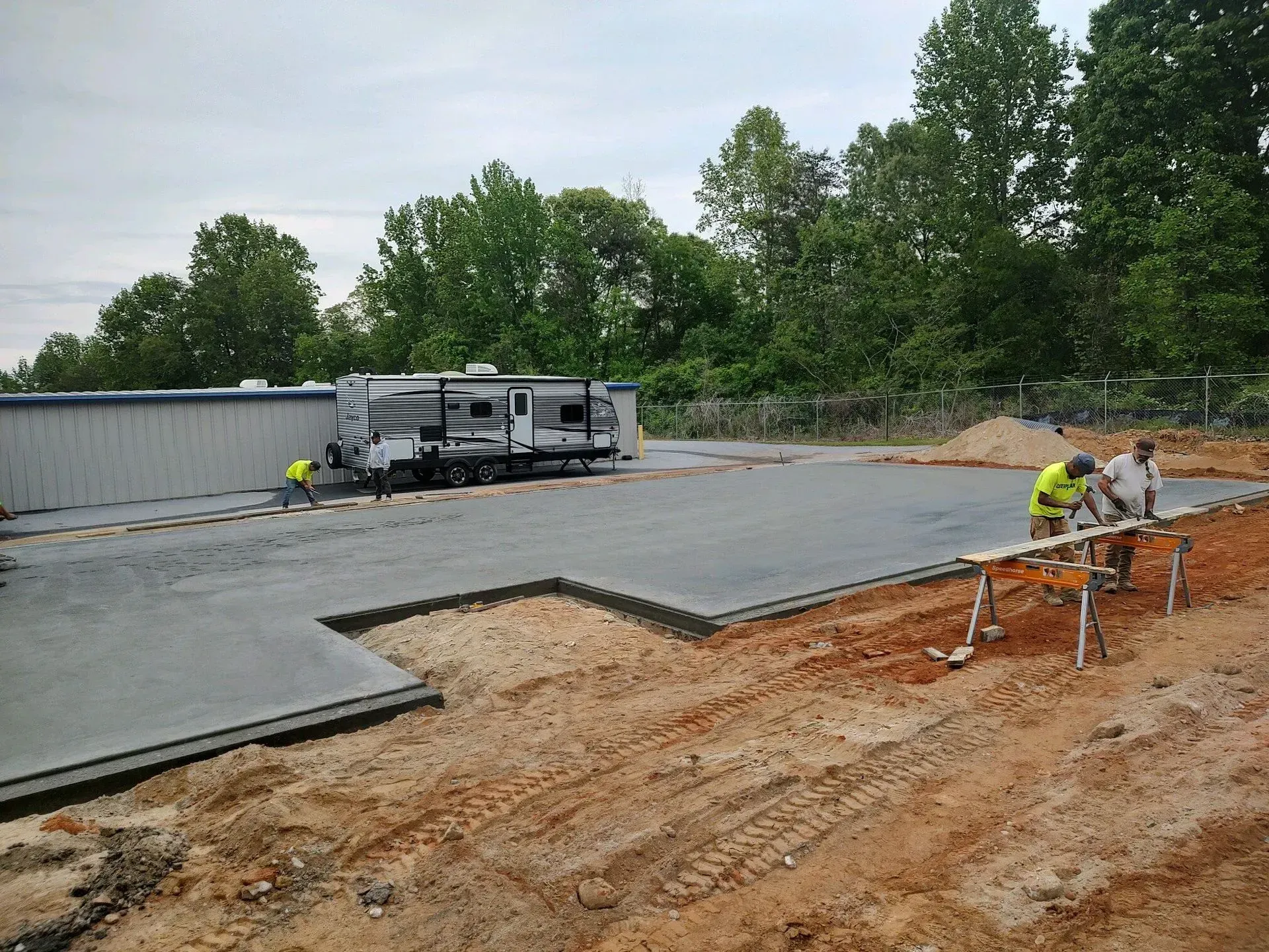 Construction crew pouring a concrete slab beside a building under construction, with equipment and trees nearby