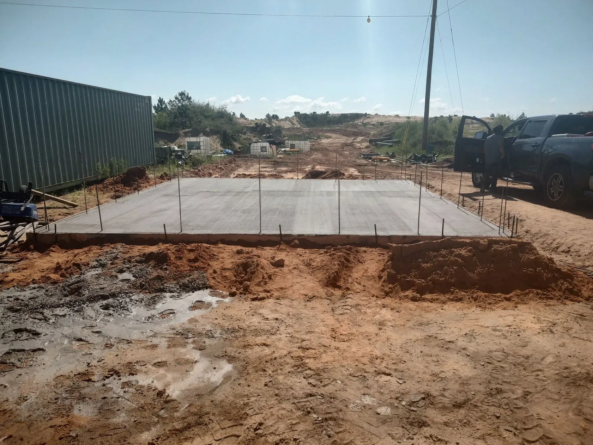 Concrete slab under construction beside a dirt lot, with fencing and a truck on the right.