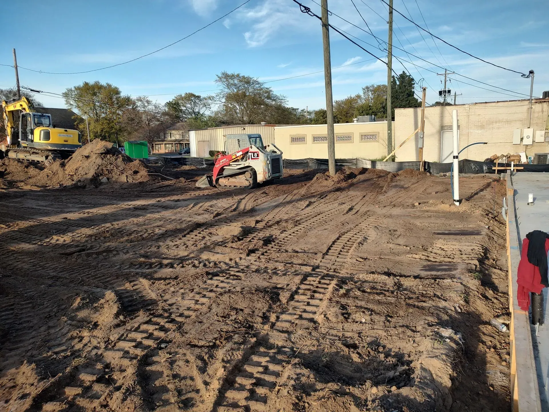 Construction site with muddy ground, a small skid steer, excavator, and trailer under a clear sky.