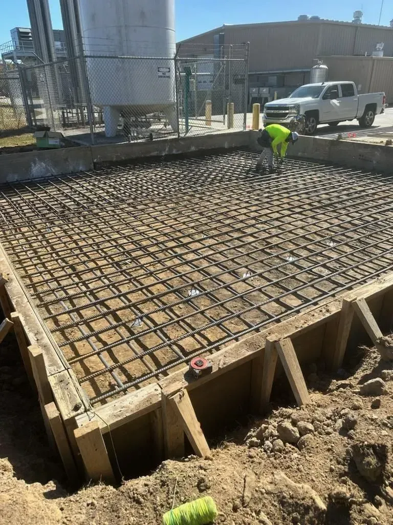 Construction site with rebar grid over formwork beside a worker in a neon shirt and a white truck