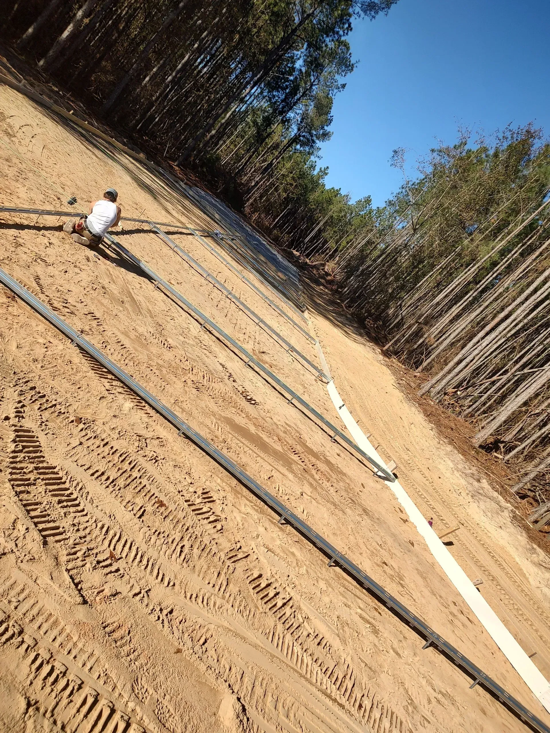 Construction site with sandy ground, white pipes laid out, and trees under a clear blue sky.