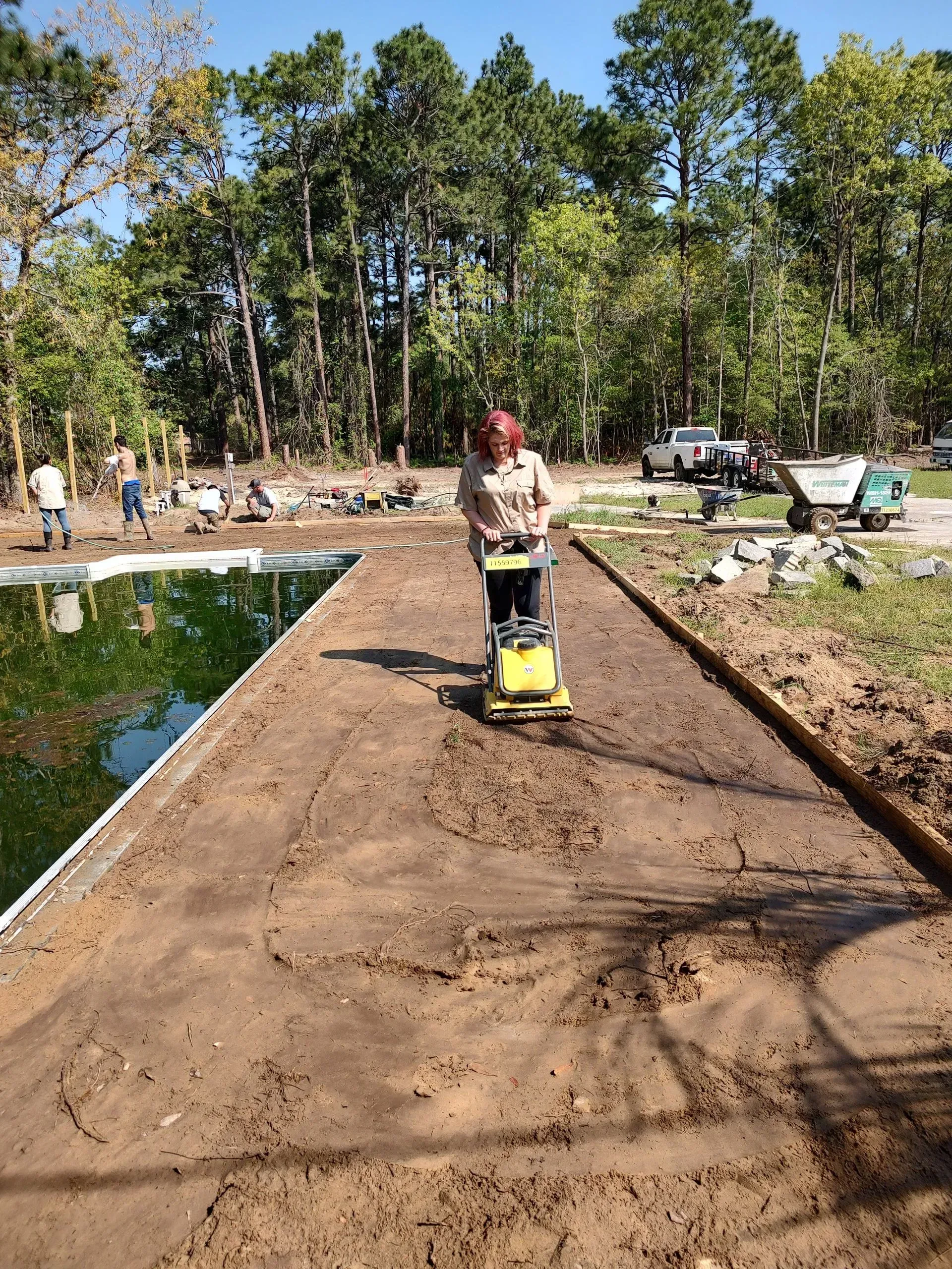 Worker operating a yellow plate compactor on a gravel path by a pond and trees