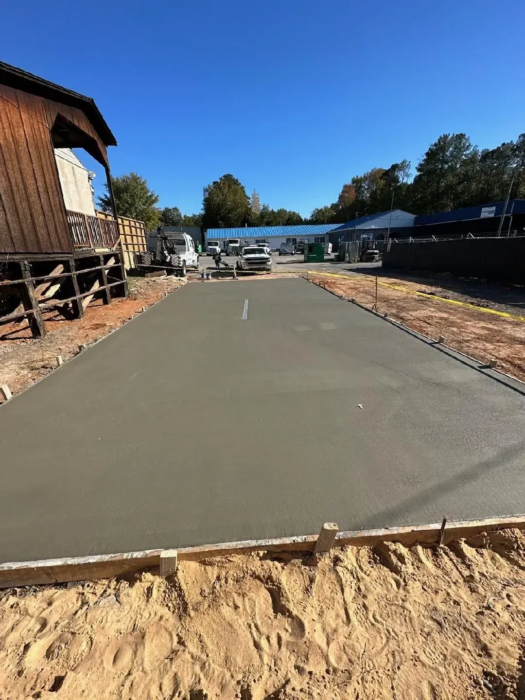Freshly paved concrete walkway at a construction site beside a wooden building under a clear blue sky