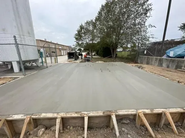 Freshly poured concrete road section at an industrial site, framed by wooden formwork and nearby trees.