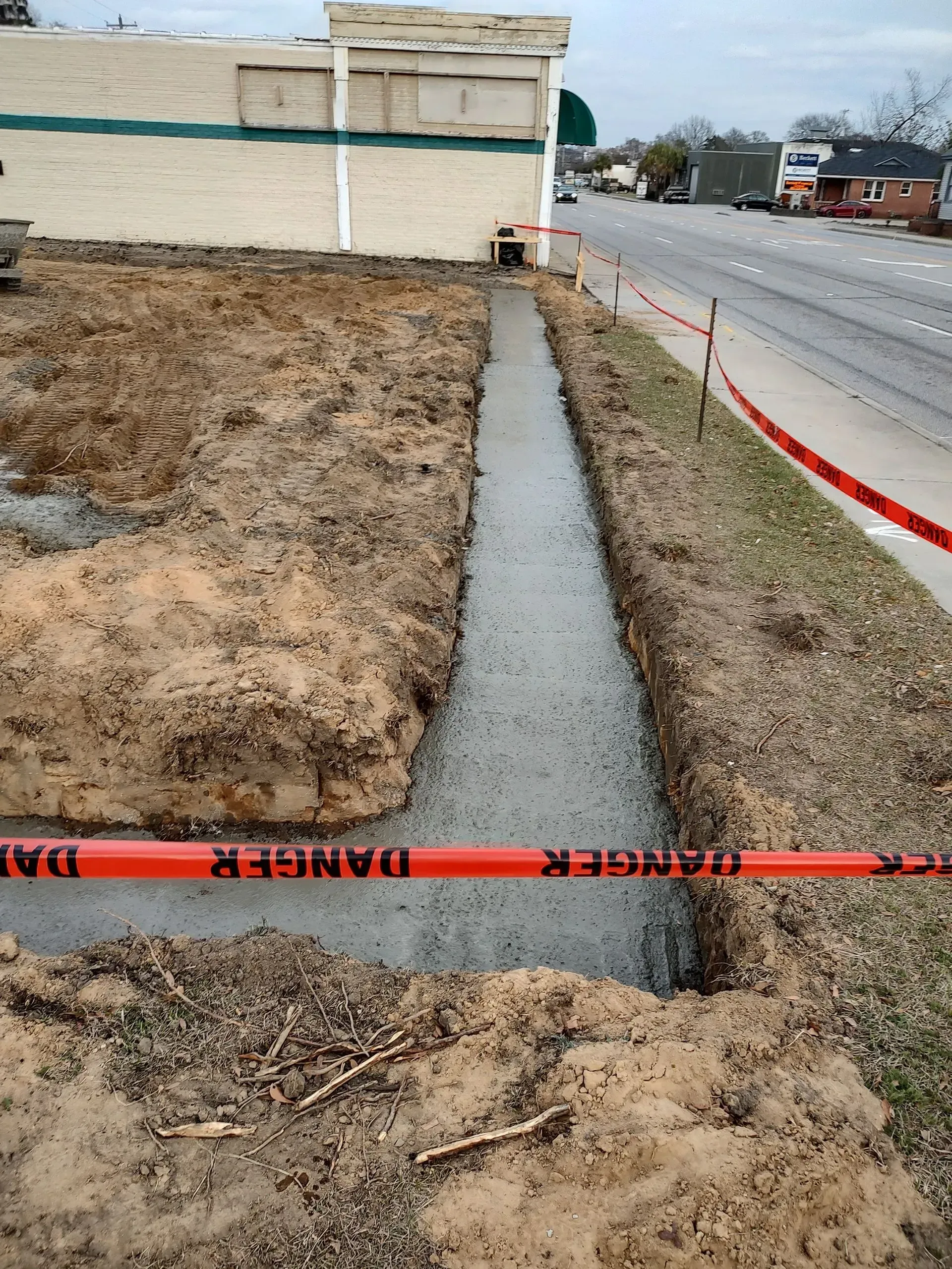 Open trench along a roadside sidewalk, marked with orange safety tape near a grassy lot.