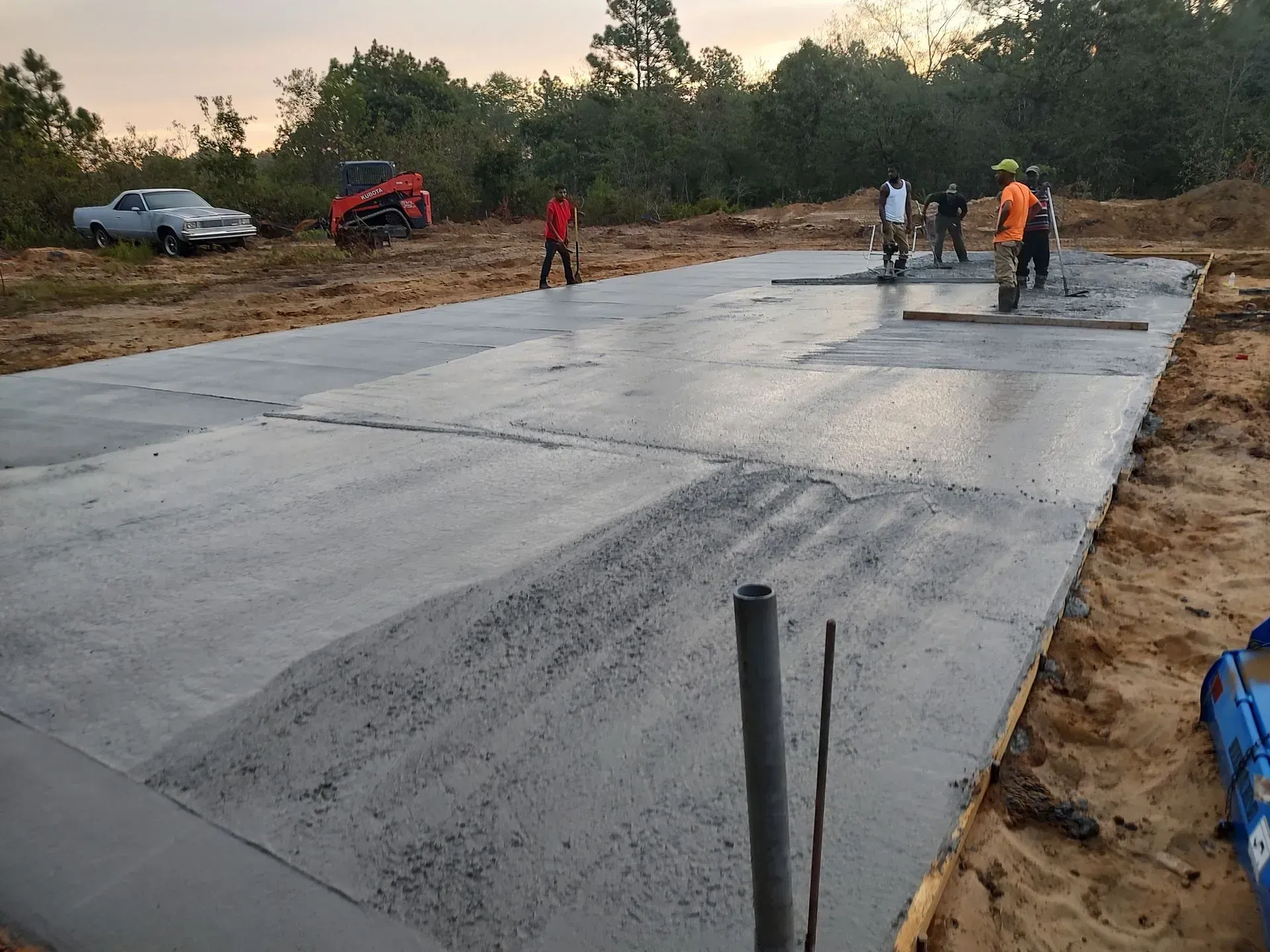 Workers smoothing wet concrete on a large driveway slab outdoors, with vehicles and trees nearby.