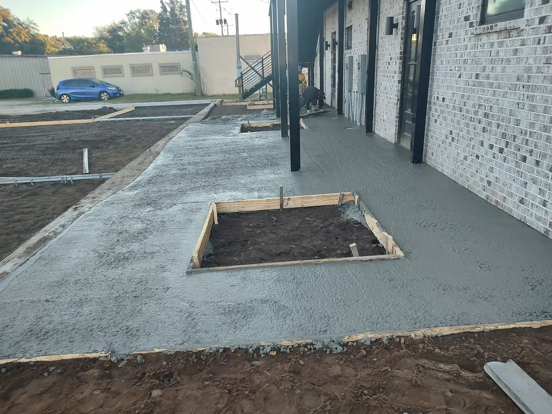 Concrete walkway beside building with a square tree pit in the foreground and a gravel path beyond.