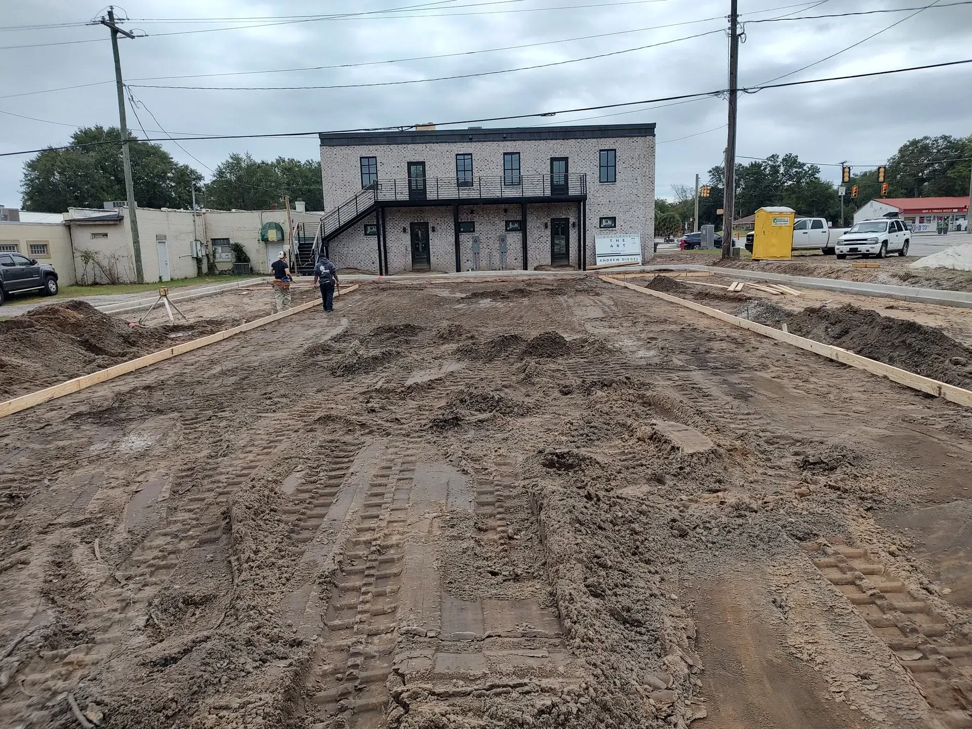 Muddy construction site leading to a two-story gray building, with one person walking and vehicles in the background