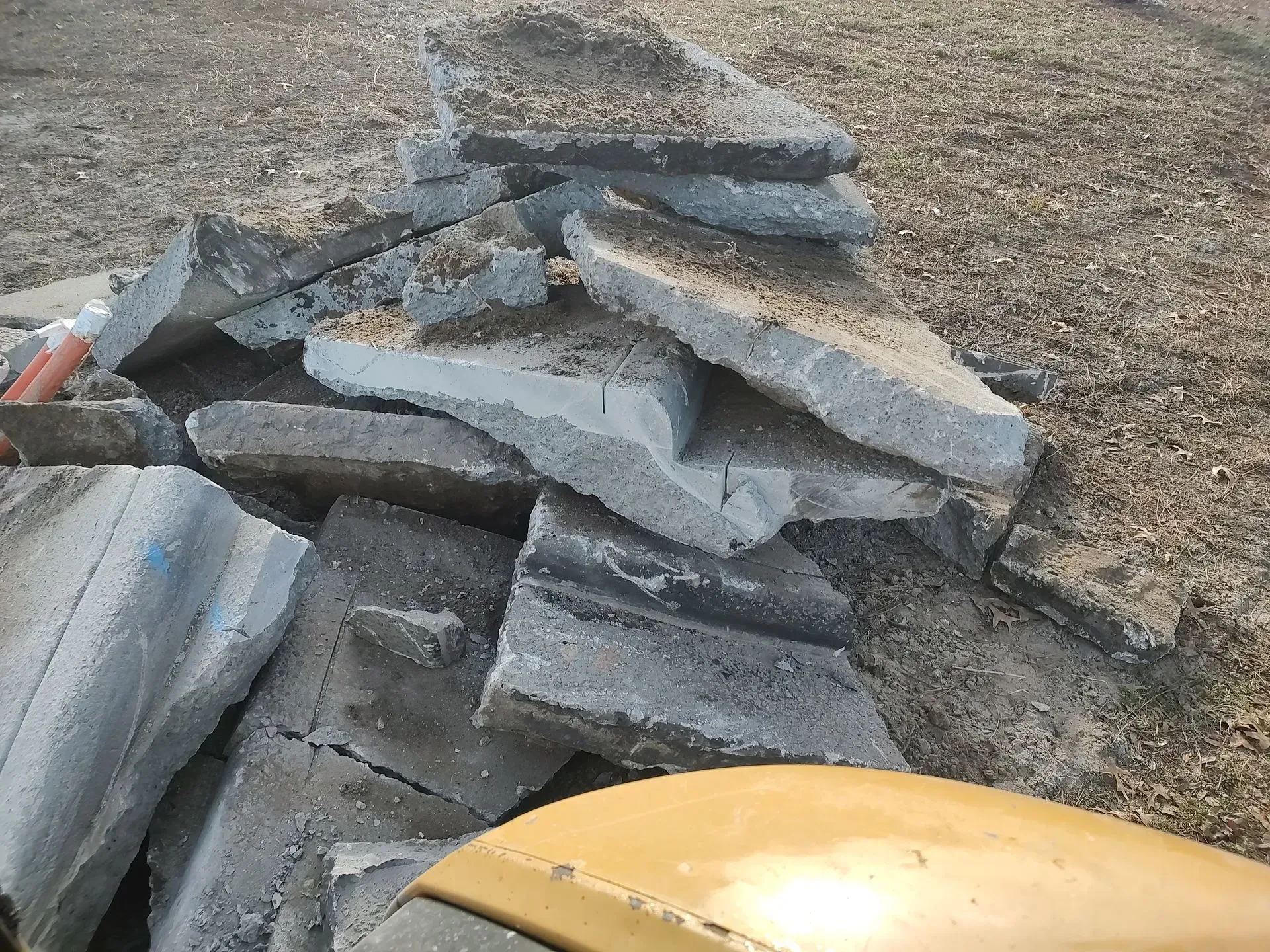 Stacked broken gray concrete slabs on dirt beside a yellow vehicle hood