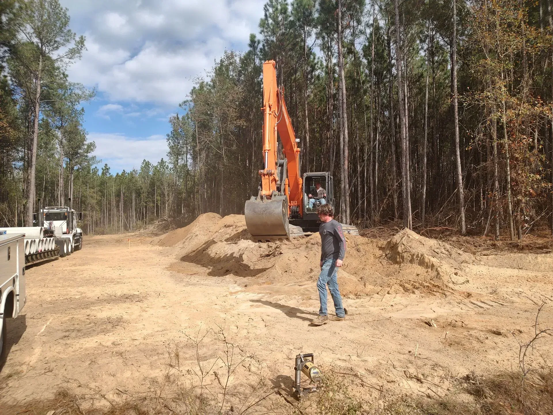 Excavator and worker on a dirt road clearing land beside a forest under a partly cloudy sky