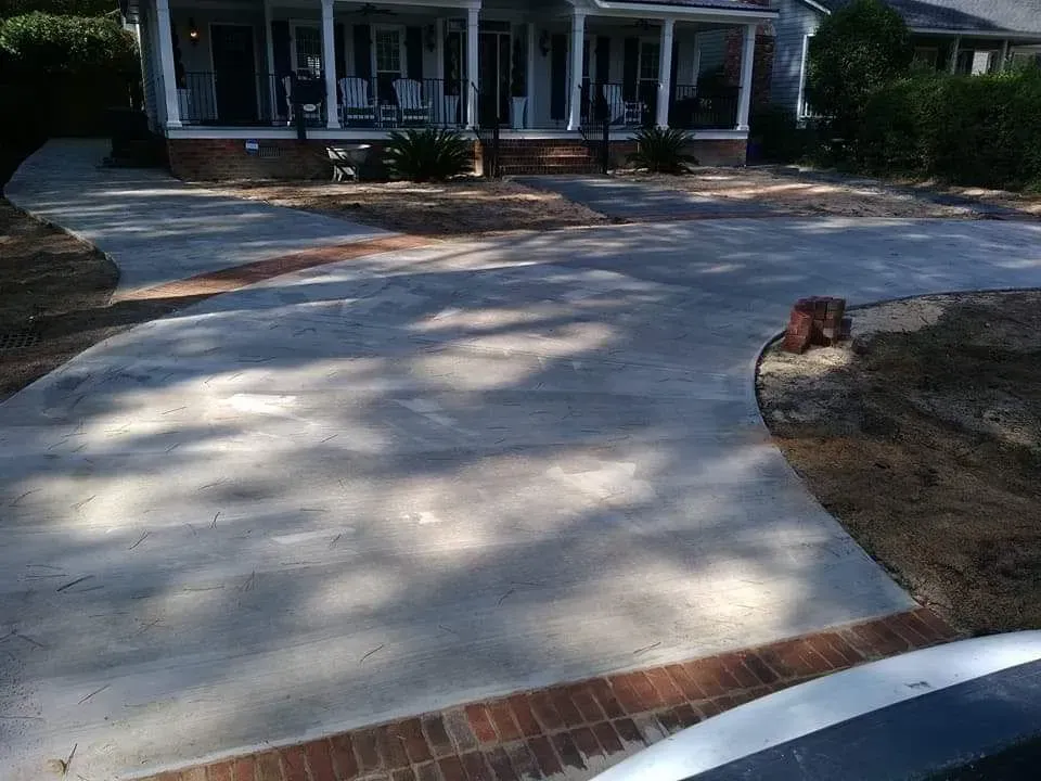 Curved concrete driveway leading to a house with a front porch, brick edging, and tree shadows.