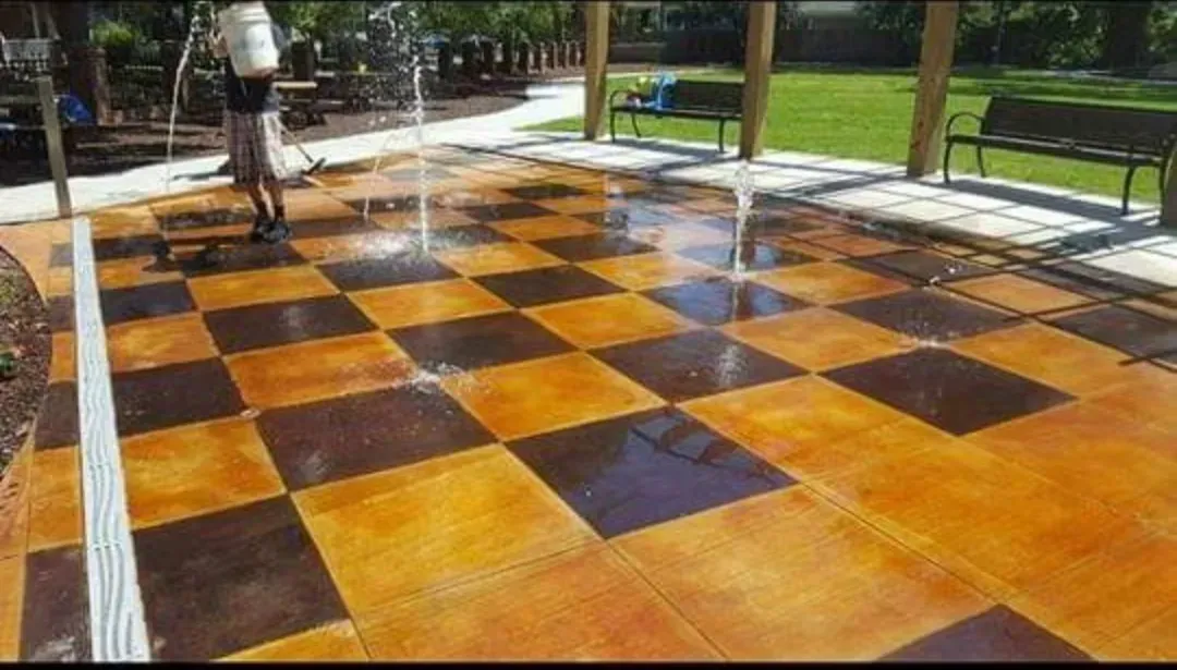 Wet orange-and-black checkerboard patio tiles with water spraying across them in a park.