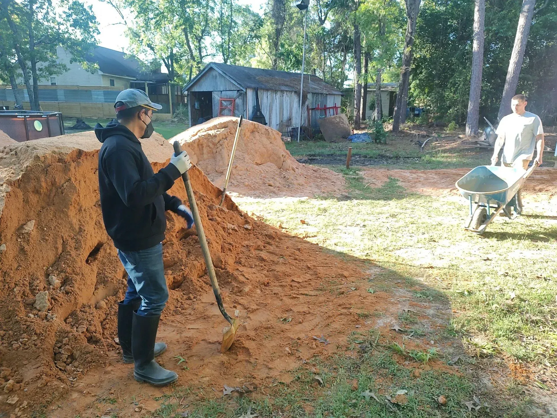 Two workers moving soil with shovels and a wheelbarrow in a sunny yard with houses nearby