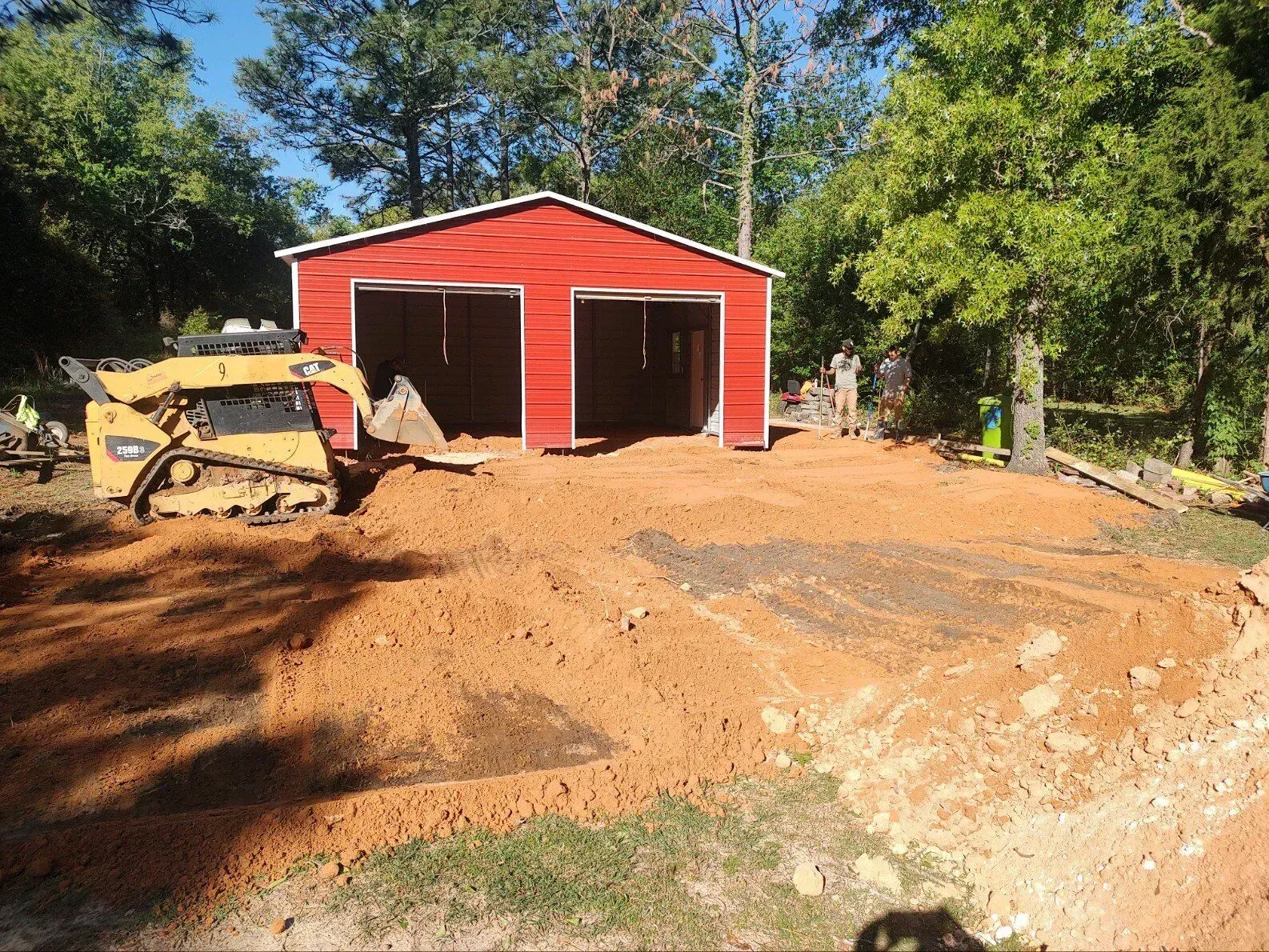 Red garage under construction with a bulldozer and dirt pile in the foreground, trees around it.