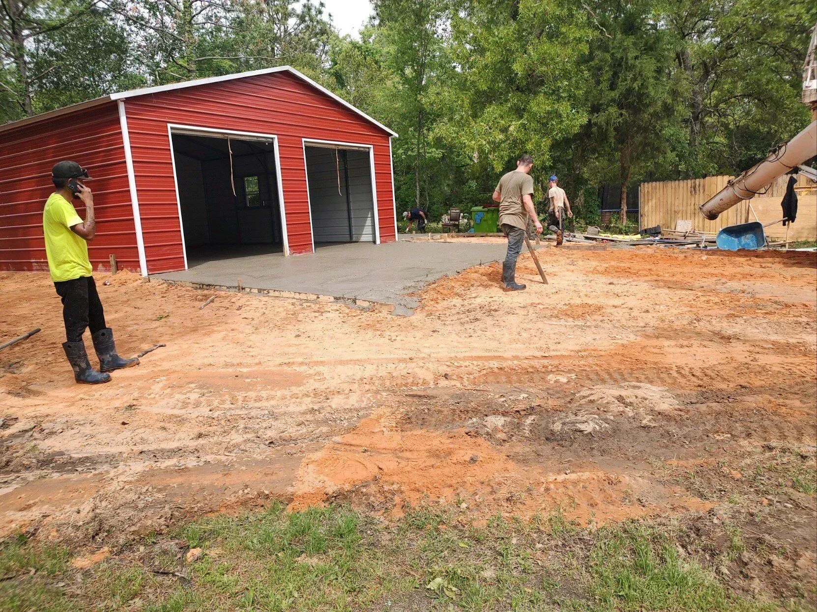 Workers spread gravel beside a red garage in a wooded yard