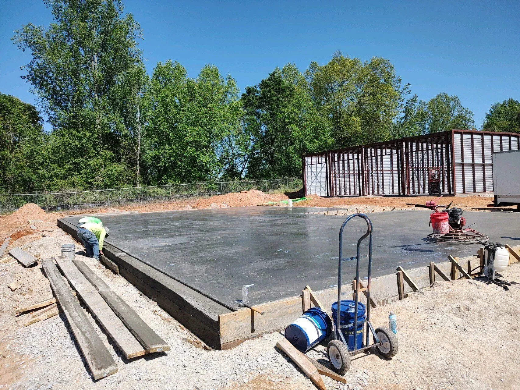 Concrete slab being poured at a construction site with workers, tools, and lumber beside a fenced structure