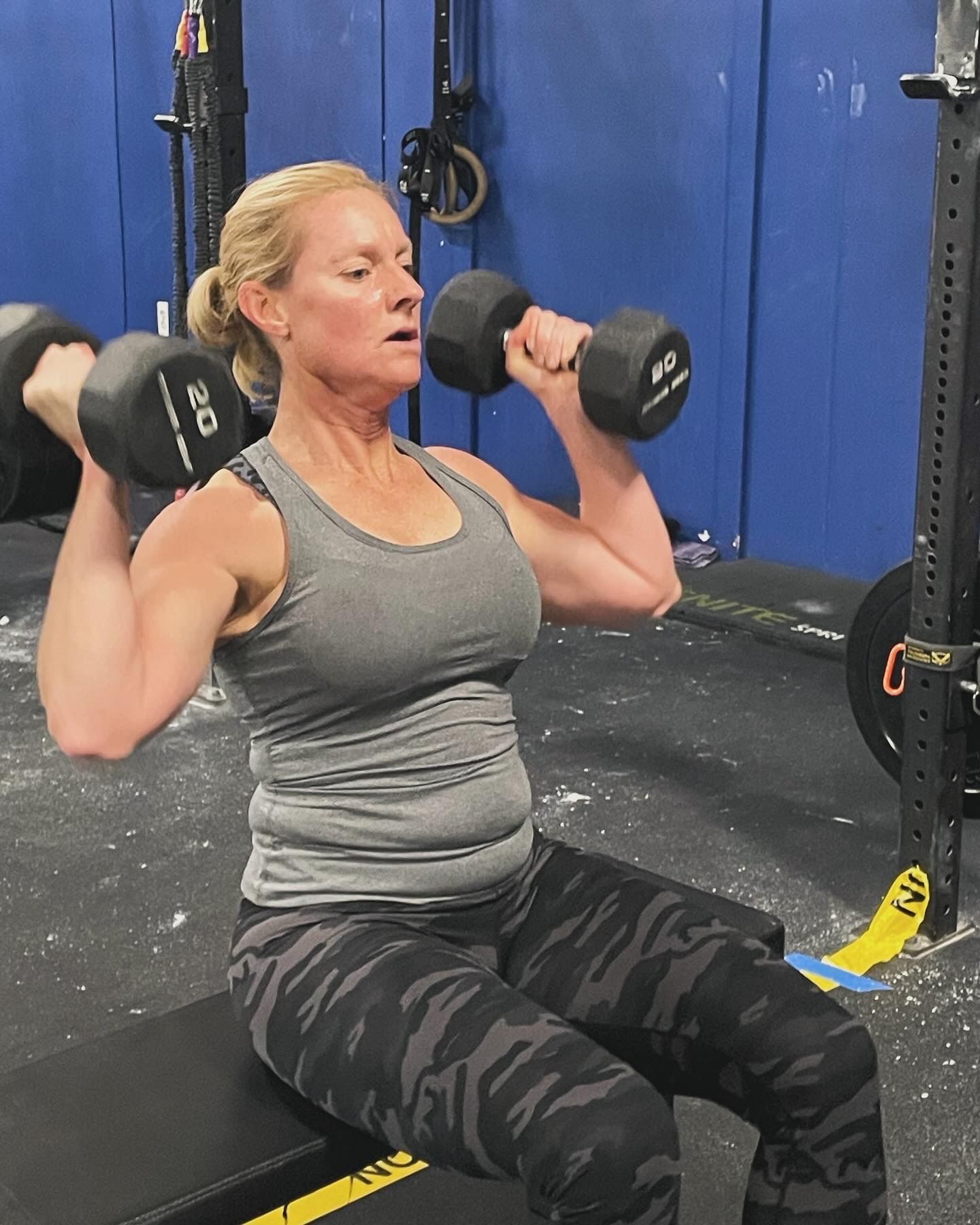 A woman is sitting on a bench lifting dumbbells in a gym.