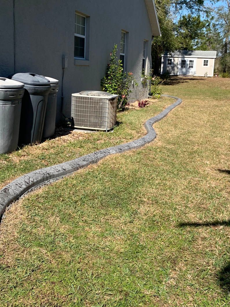 A gray concrete edging snakes across a grassy yard, near a house and AC unit.