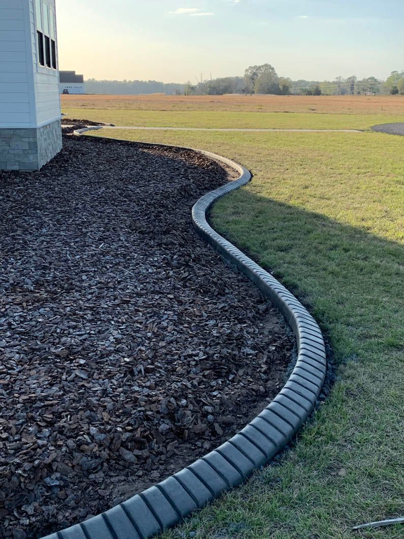 Dark gray concrete edging separates brown mulch bed from green grass lawn next to a white house.