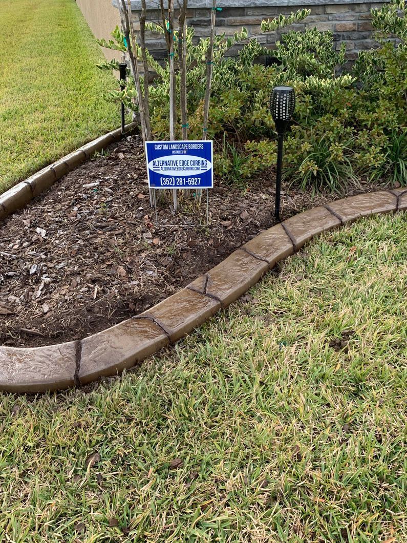 Stone-like concrete edging borders a garden bed filled with mulch and plants, adjacent to a green lawn.