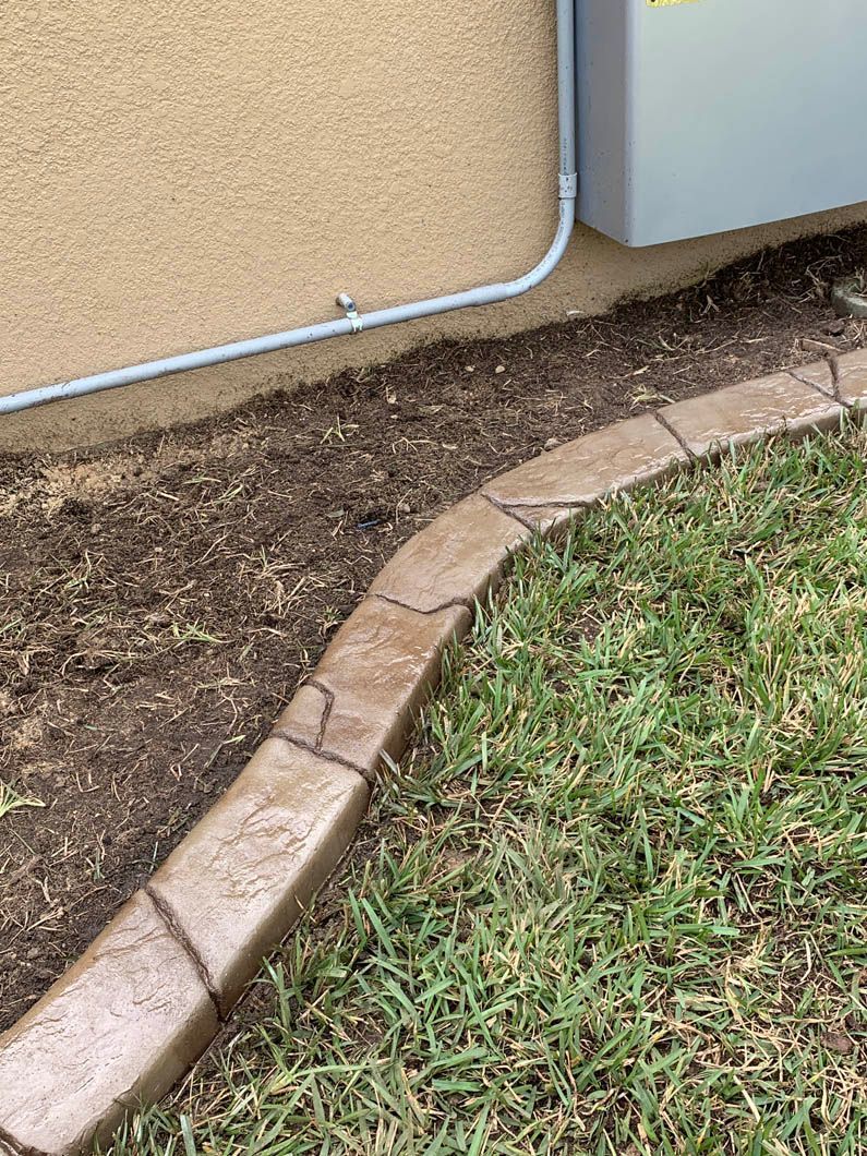 Brown stone-like concrete edging separates dirt from grass, adjacent to a tan stucco wall and metal box.