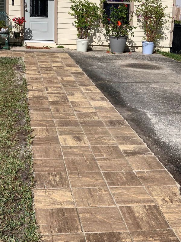 Brown tile walkway leading to a beige house with potted plants. A driveway is to the right.