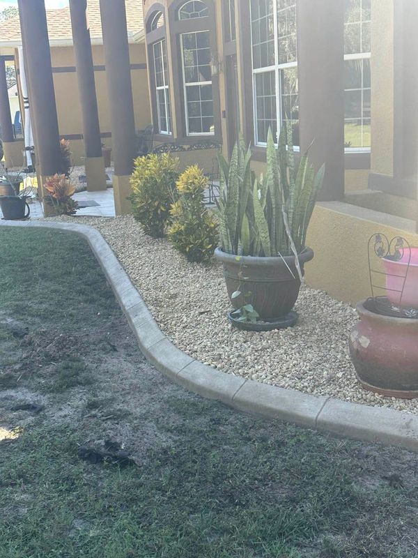 Concrete-bordered landscaping bed with rock, plants, and large pot against a light-colored house.